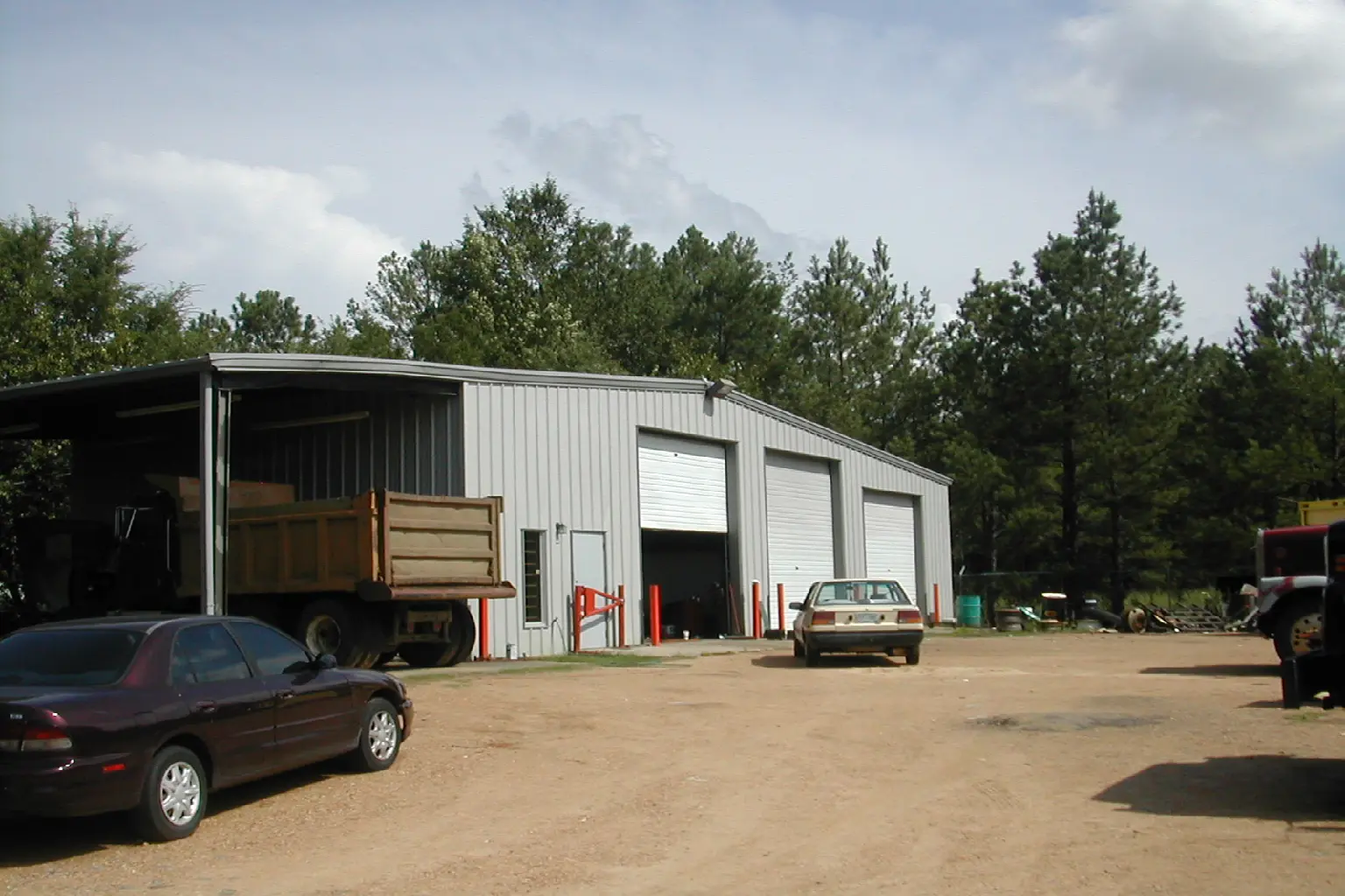 A metal warehouse with open garage doors and vehicles outside on a dirt lot.